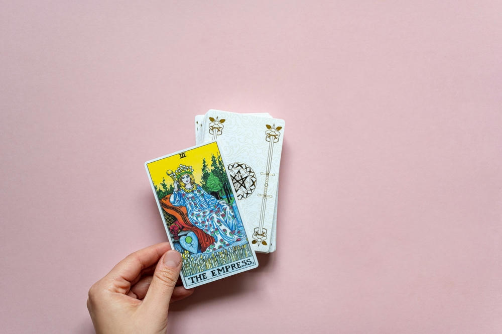 The Empress Tarot Card of Rider Waite deck in hand on a pink background.
